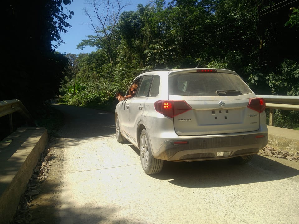 Yvonne zwaait uit het raam van een stoffige zilveren Suzuki Vitara op een brug langs een regenwoudweg in Costa Rica.