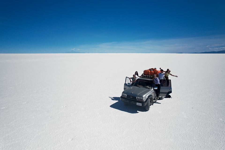 Een Toyota Land Cruiser met een oranje dakbagagetas op de witte zoutvlakte van Bolivia, met zwaaiende reizigers in de deuren.