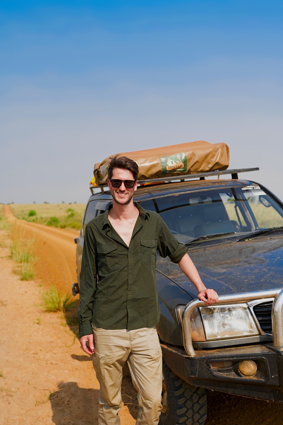 Martijn leunt op de bullbar van een 4x4 met daktent, naast een rode zandweg door de graslanden van Oeganda.